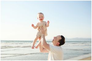 Father joyfully lifting laughing baby high in the air on a sunny beach with ocean waves in the background.