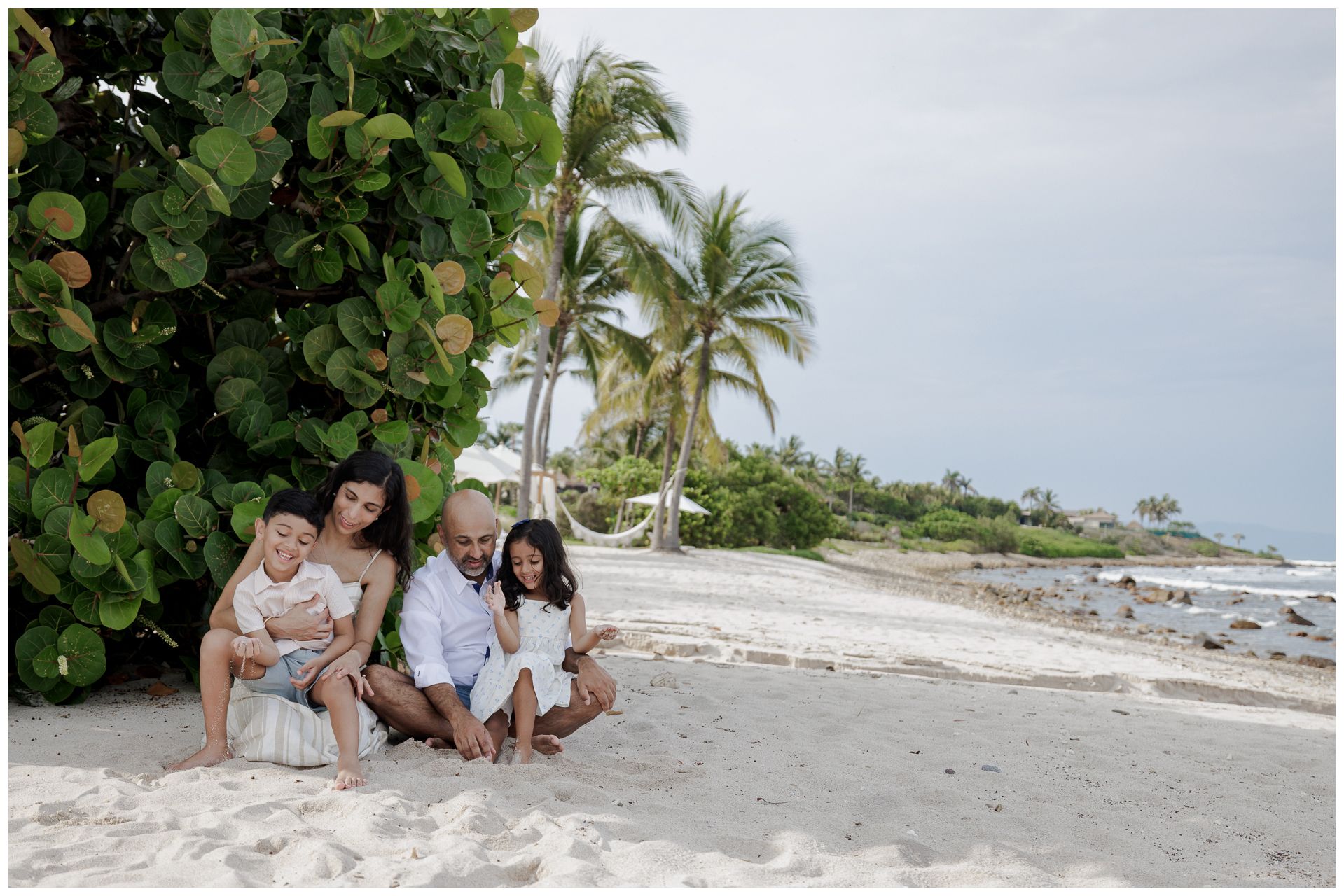 Family of four sitting together on a white sand beach near palm trees and tropical foliage.