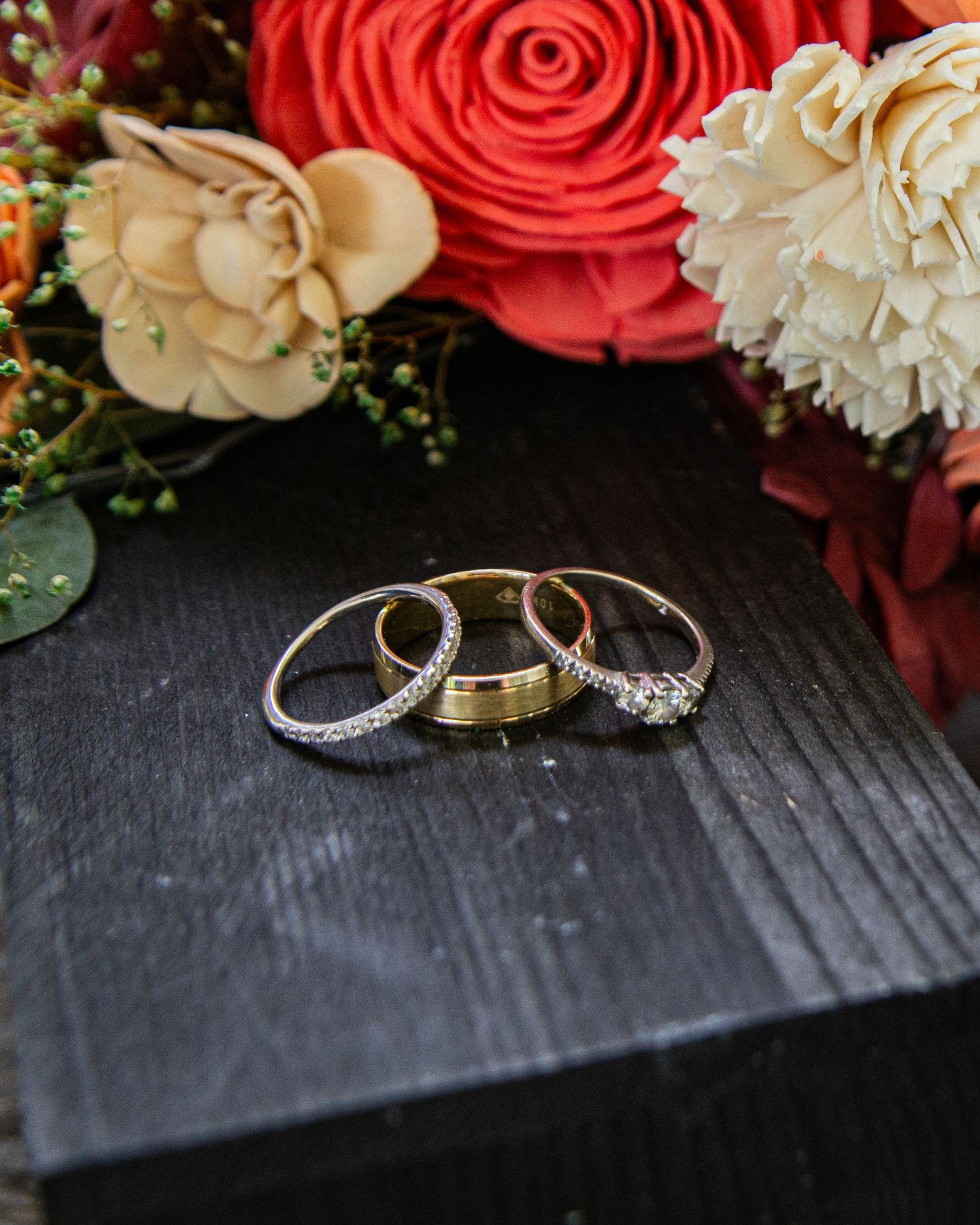 Three wedding rings stacked on dark wood surface with red and cream floral bouquet in background.