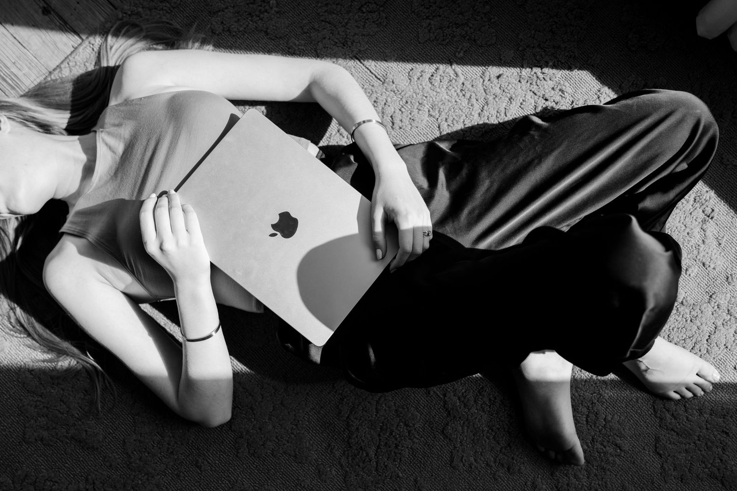 Black and white photo of two people lying on floor with Apple MacBook laptop between them.