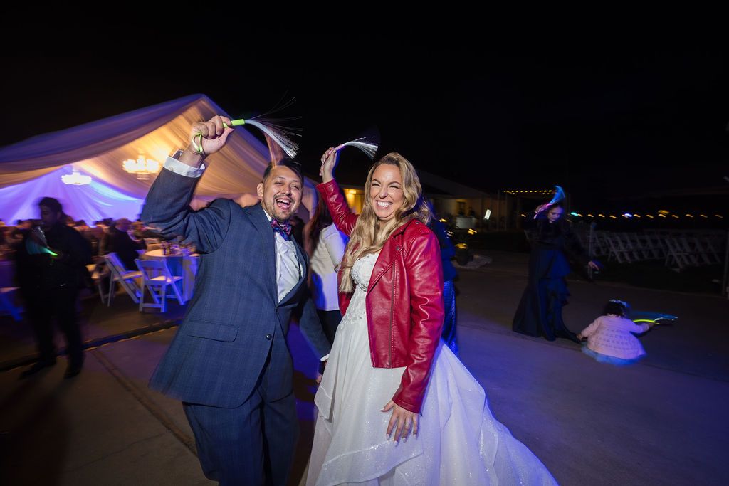 Bride in white gown and red leather jacket dances with groom in blue suit at outdoor wedding reception at night.
