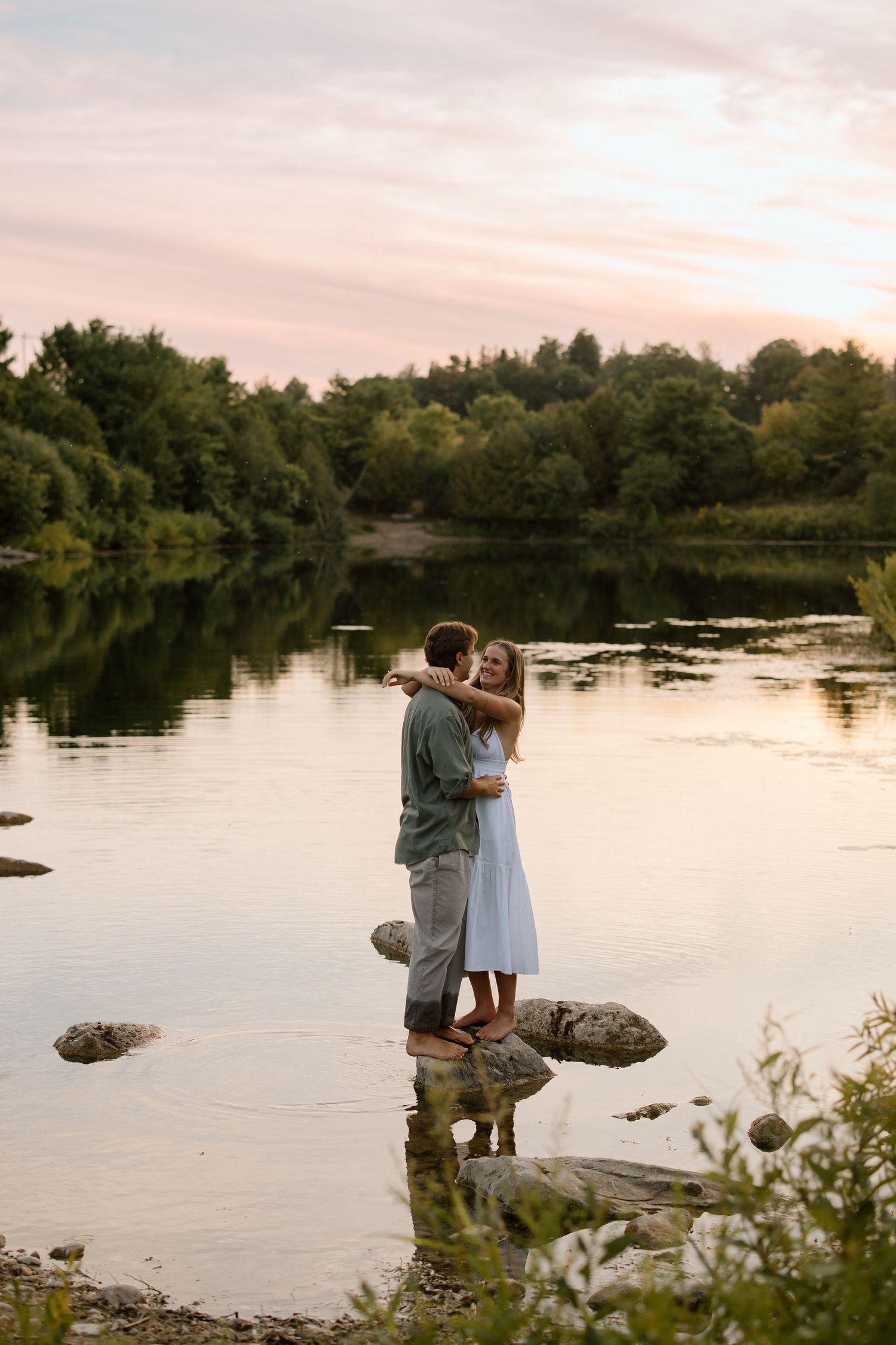 Couple embracing on rocks by a serene lake at sunset, surrounded by lush green trees reflecting on calm water.