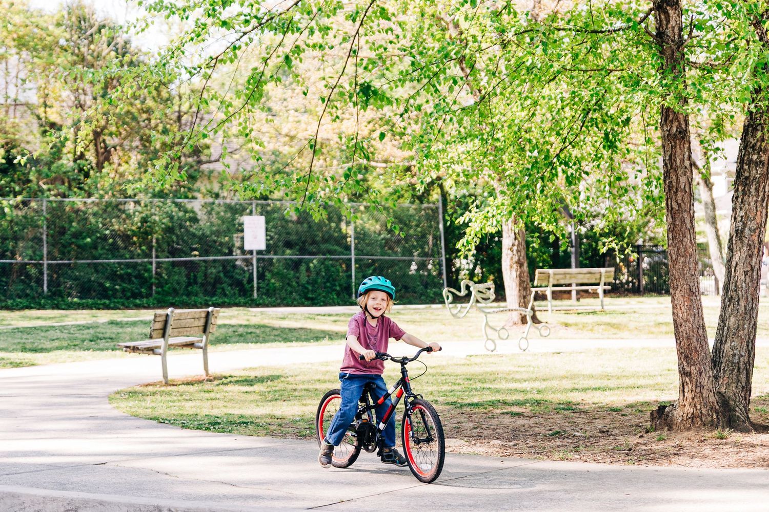 Child wearing a helmet rides a bicycle on a paved path in a sunny park with green trees and benches.