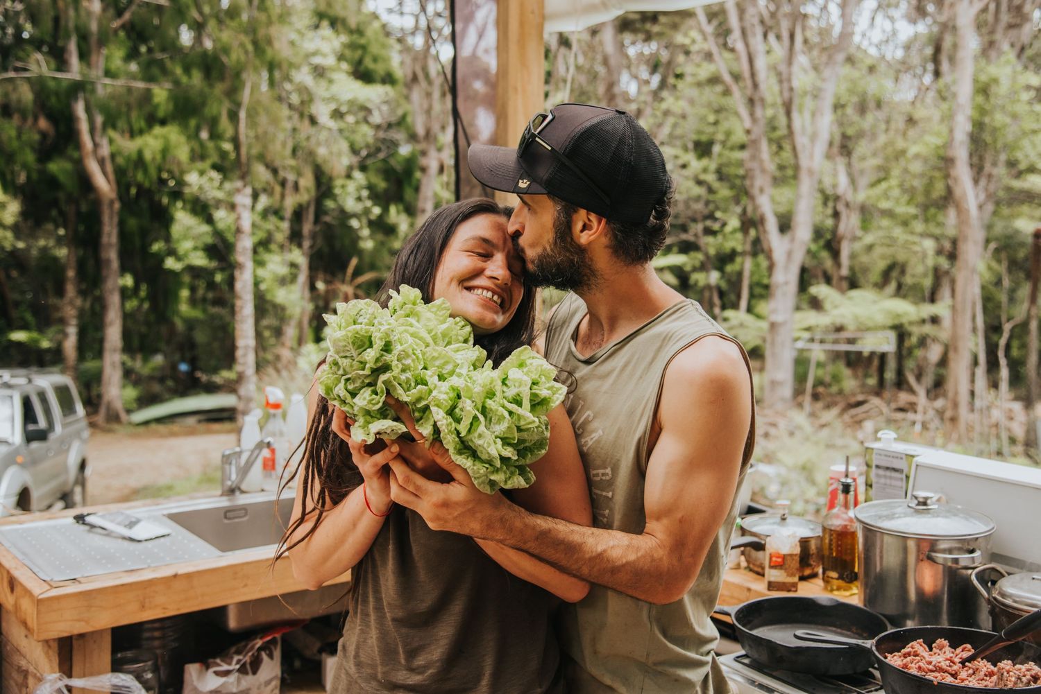 A couple in white and pink attire poses joyfully in a green outdoor setting on a cloudy day.