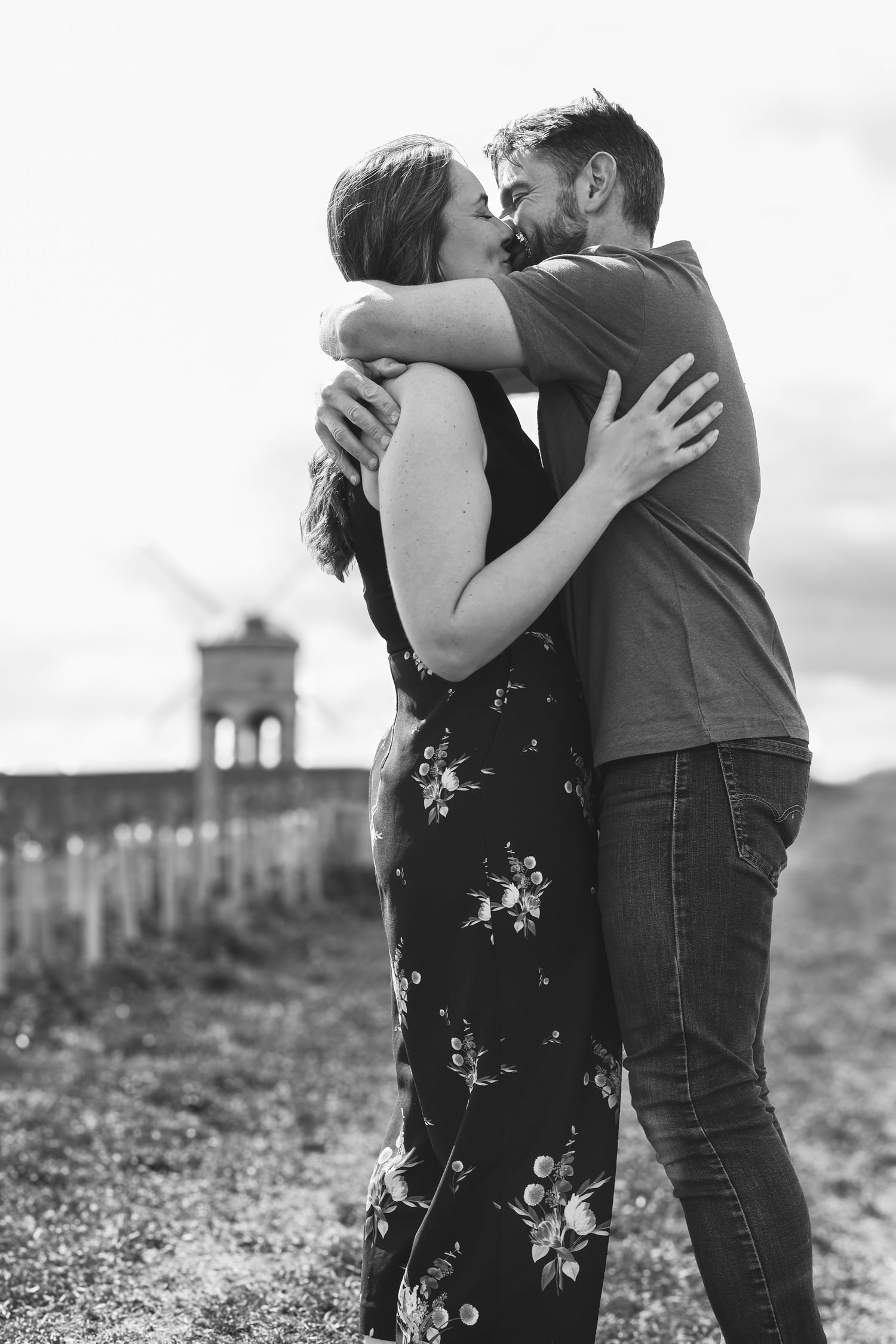 Black and white photo of a couple embracing and kissing outdoors, woman in floral dress, man in polo shirt.