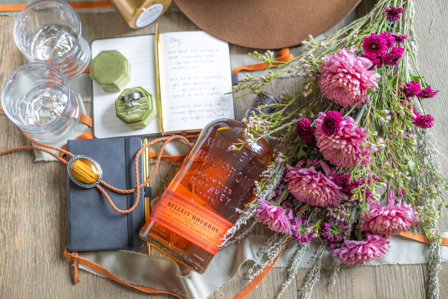 An elegant wedding flat lay featuring a bolo tie, rings in green ring boxes, pink flowers and a bottle of bourbon. Vow books are present.