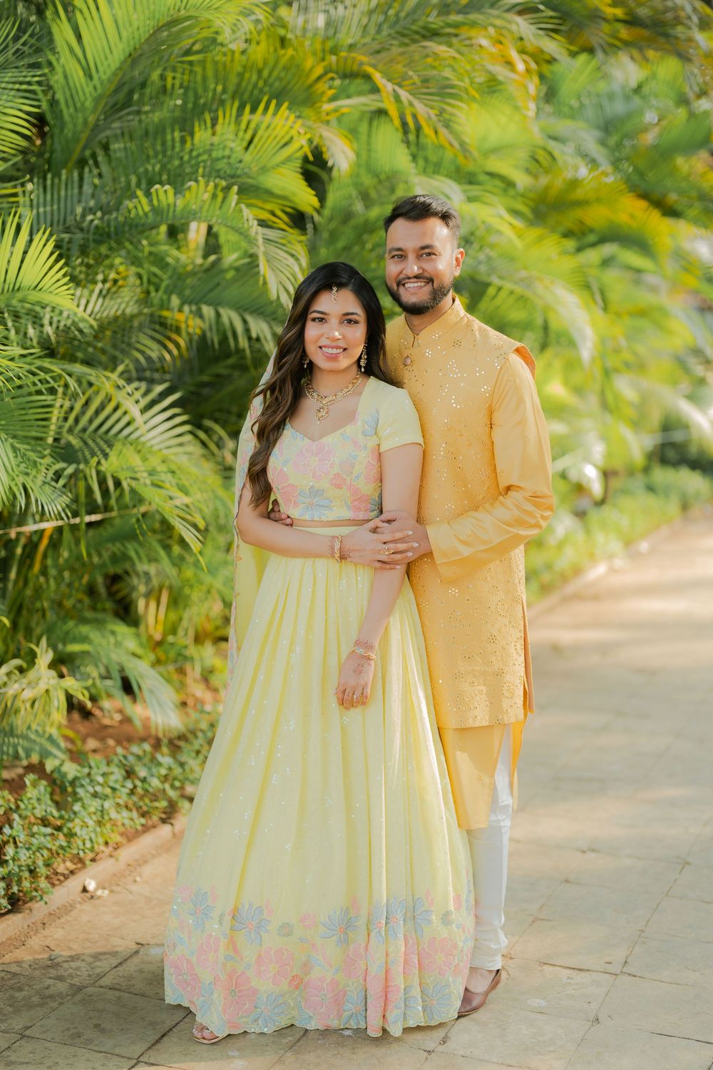 Couple in matching yellow Indian outfits posing together in a lush tropical garden setting.