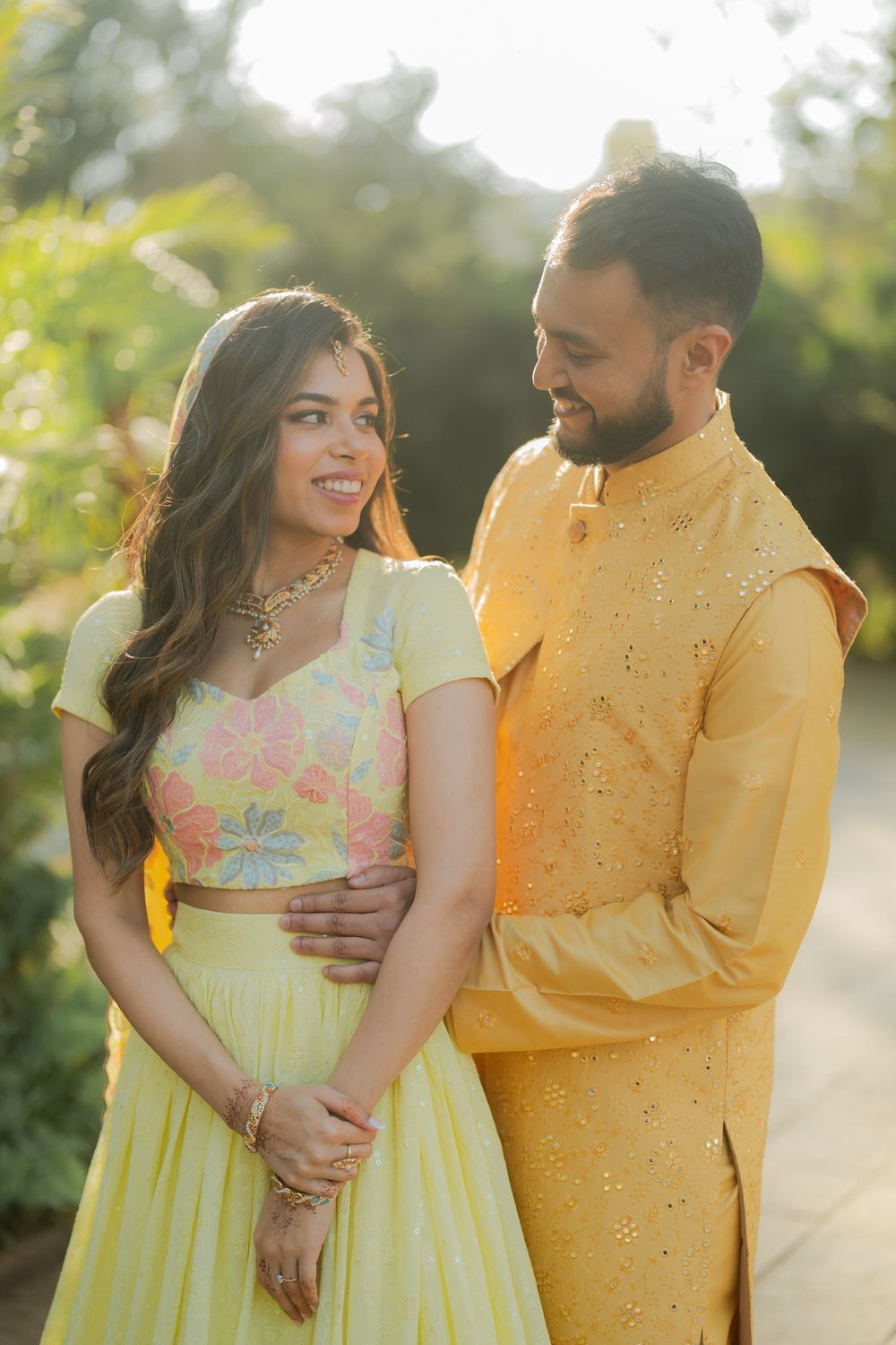 Couple in coordinating yellow Indian outfits, woman in floral lehenga and man in embroidered kurta, posing outdoors.