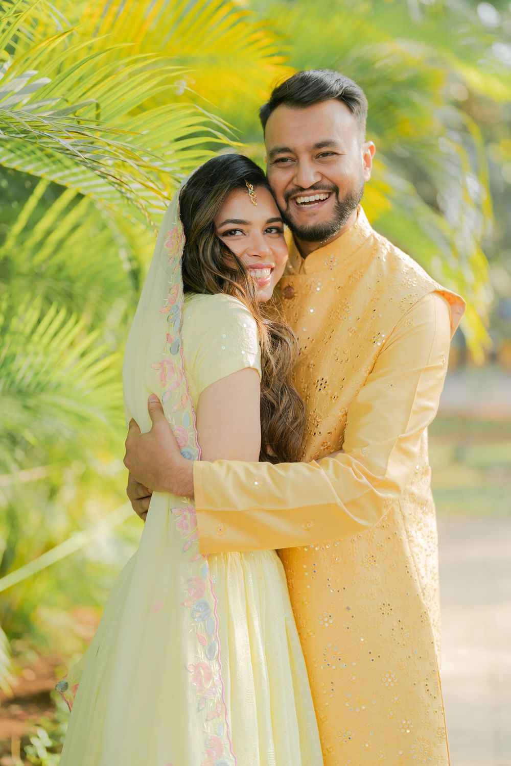 A smiling couple in matching yellow Indian outfits embracing outdoors among tropical palm trees.