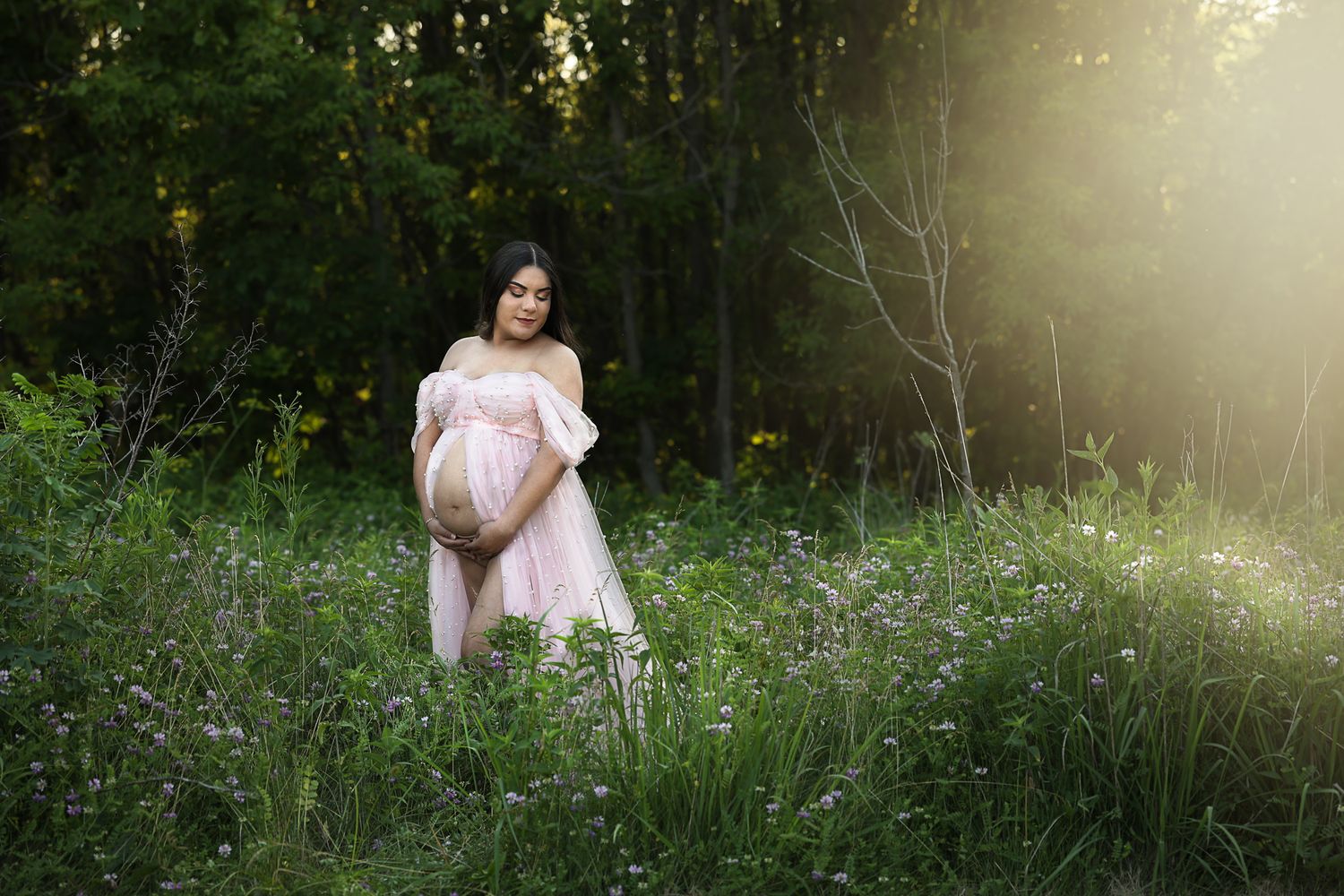Pregnant woman in pink dress standing in wildflower field with soft sunlight filtering through trees behind her.