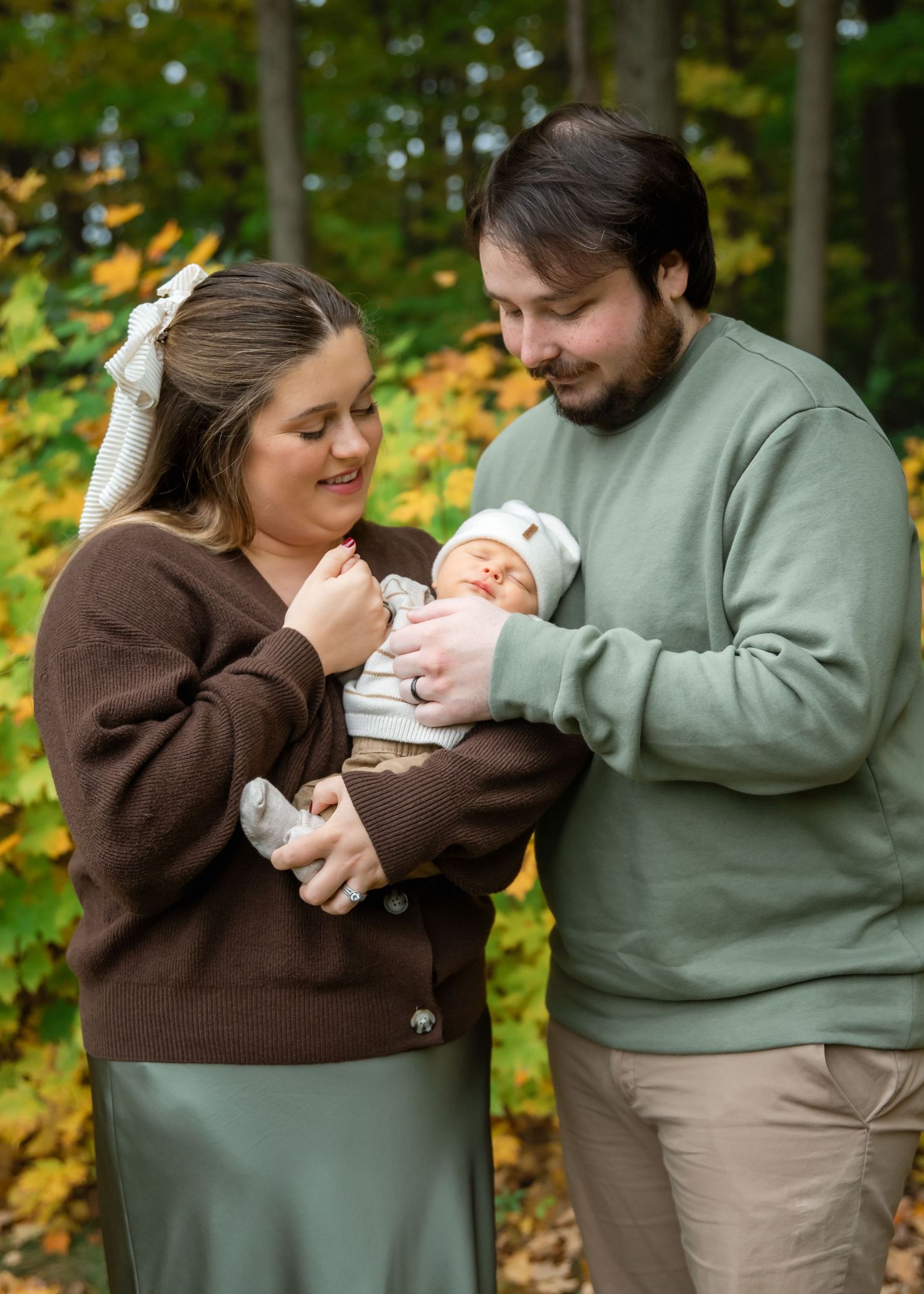 A couple in brown and green sweaters lovingly holds their newborn baby outdoors among fall foliage.