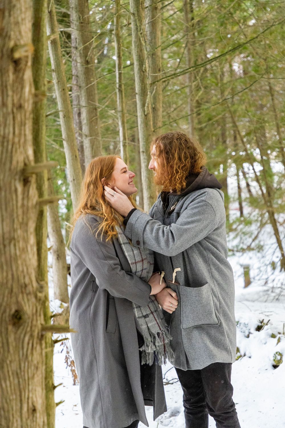 A couple in a winter forrest with tall trees. The man is fixing the woman's hair behind her ear