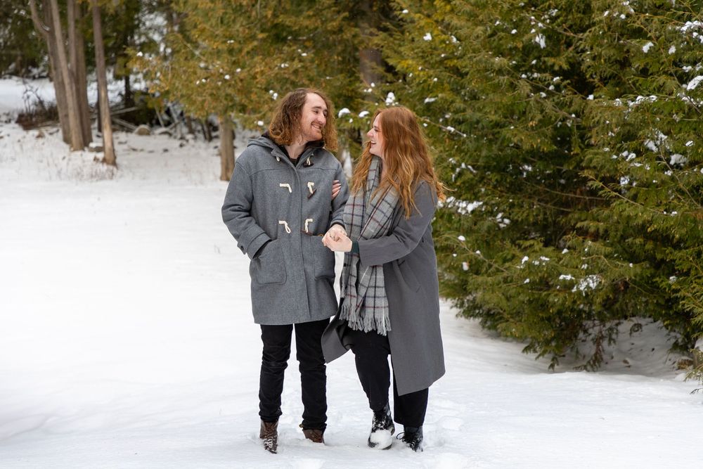 A  couple looking at each other linked arms walking through the snow with evergreen trees in the background