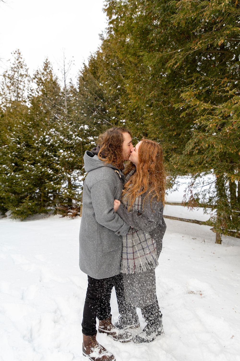 A couple kissing outside on a cold snowy day with evergreen trees behind them