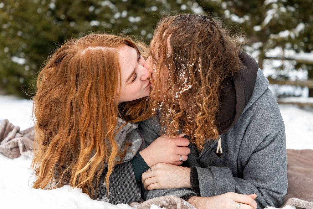 A couple cuddling in the snow on a blanket, lying down on their stomachs kissing