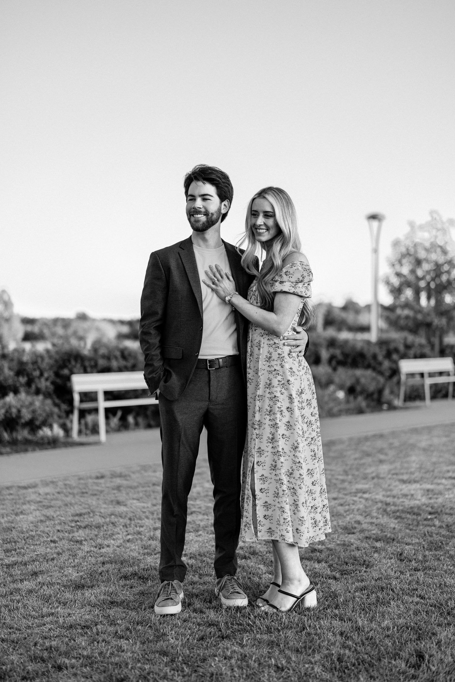 Black and white photo of a smiling couple embracing outdoors on grass, man in suit, woman in floral dress.