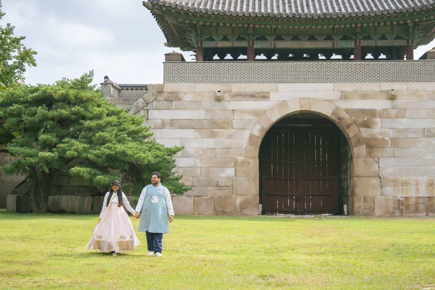 People in traditional Korean hanbok clothing walk near stone palace walls and traditional architecture.
