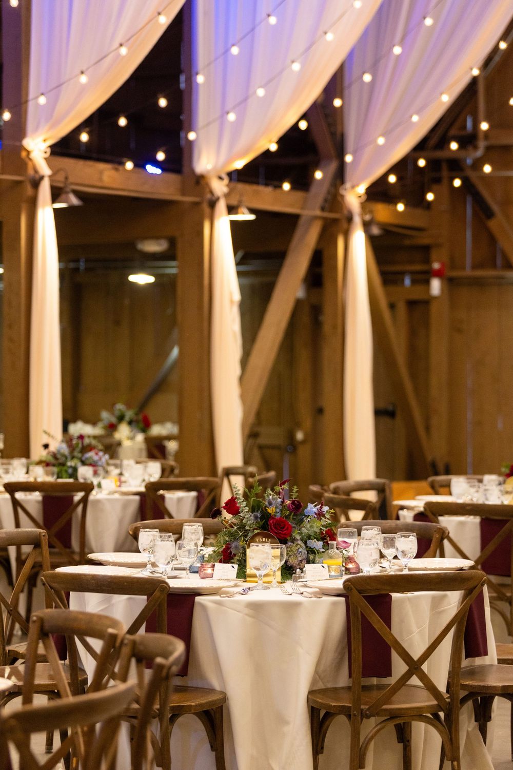 Elegant wedding reception venue with white drapes, string lights, and wooden cross-back chairs at round tables.