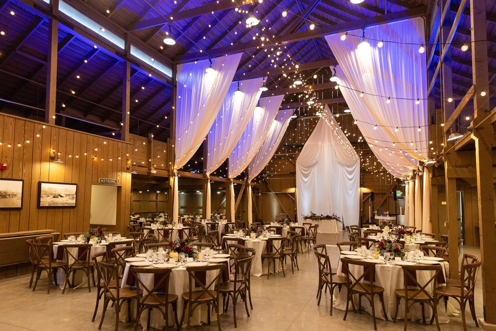Wedding reception hall decorated with white drapes, string lights, and rustic wooden chairs.