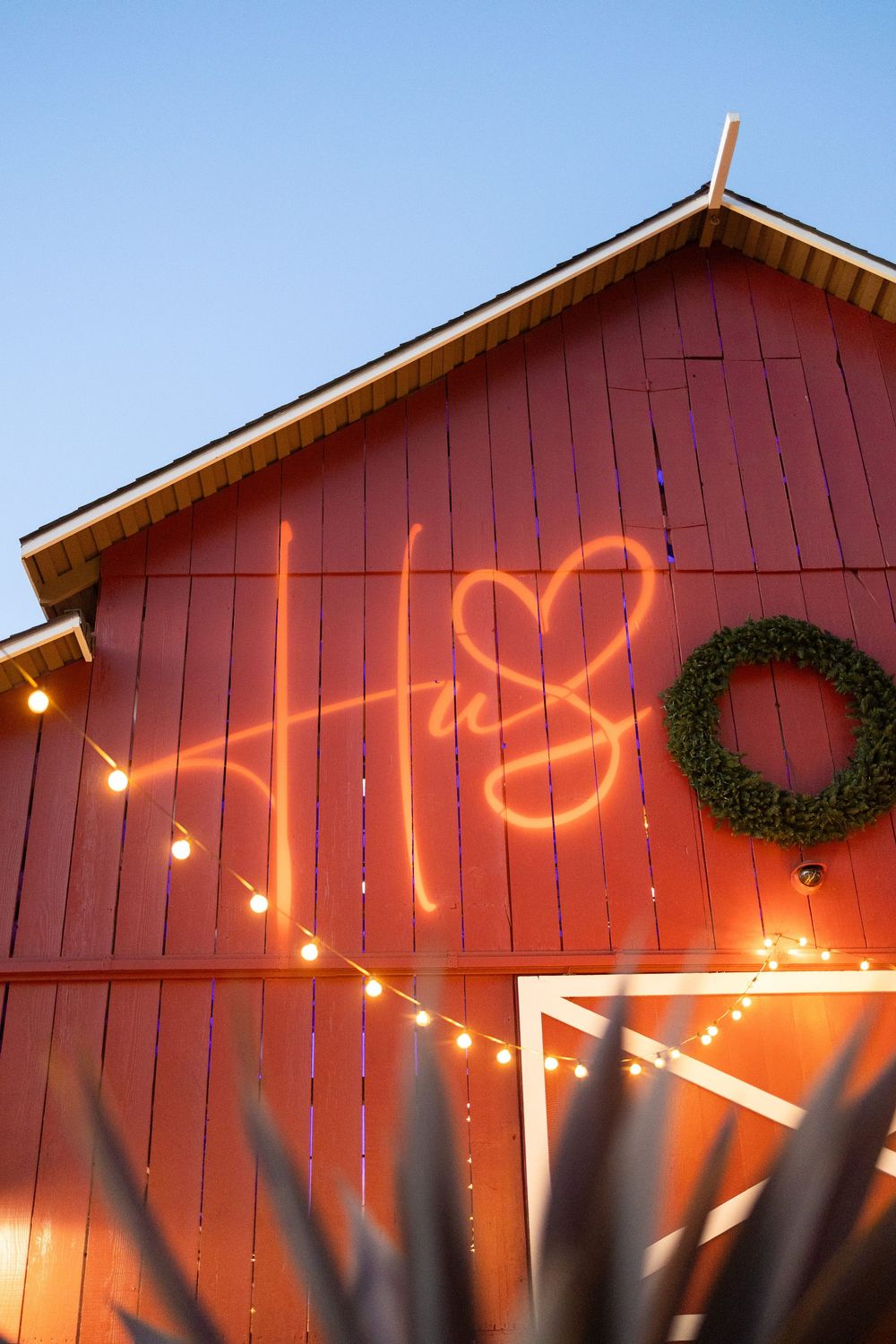 Red barn wall decorated with illuminated Christmas lights and wreaths at sunset creating a festive glow.