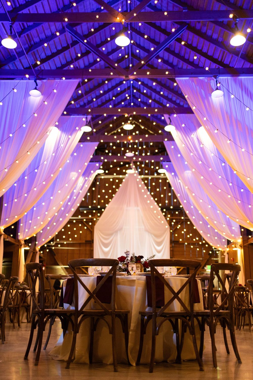Romantic wedding reception venue with draped white fabric, twinkling lights, and rustic wooden cross-back chairs.