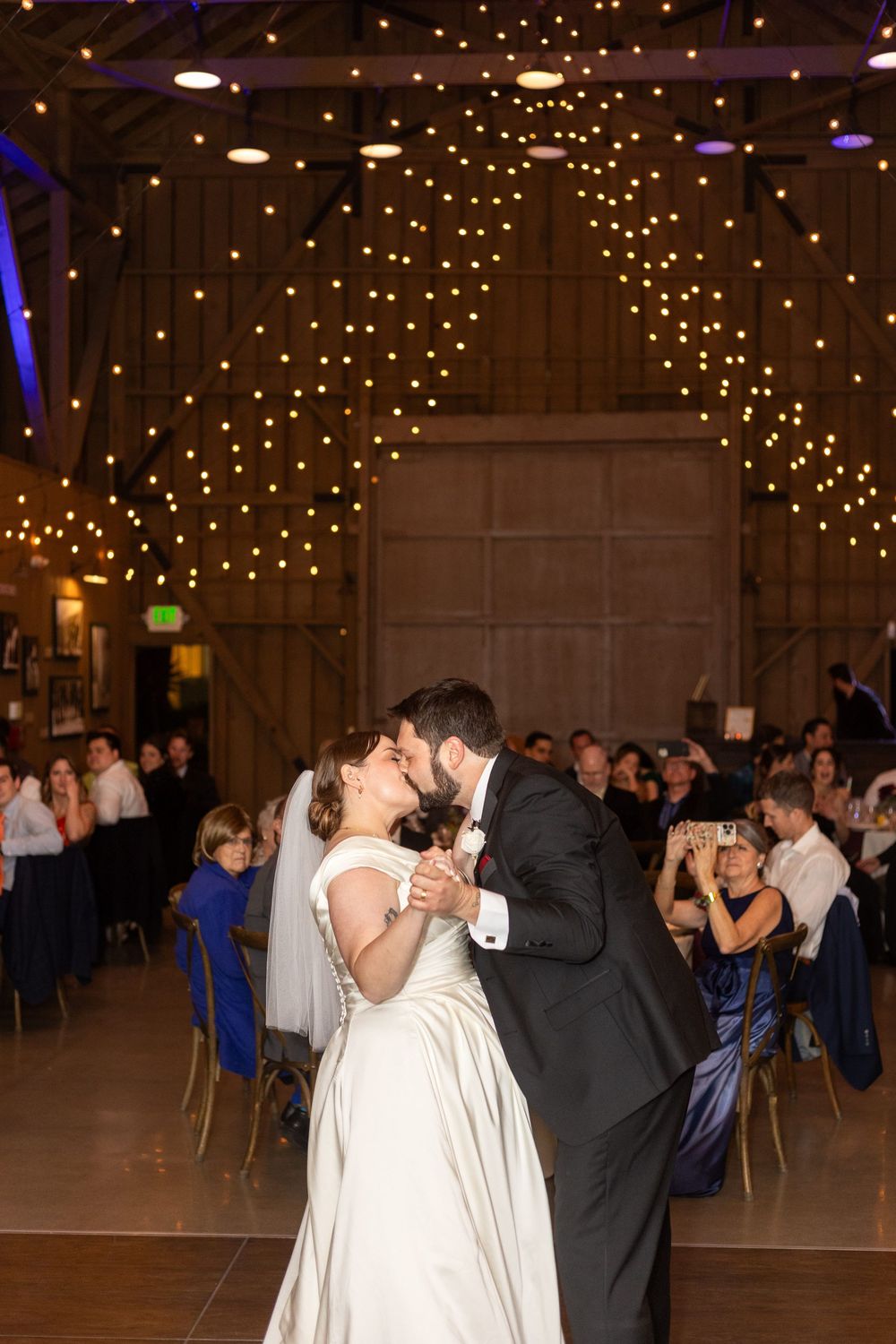 Wedding reception dance floor under twinkling string lights with couples dancing in an elegant barn venue.