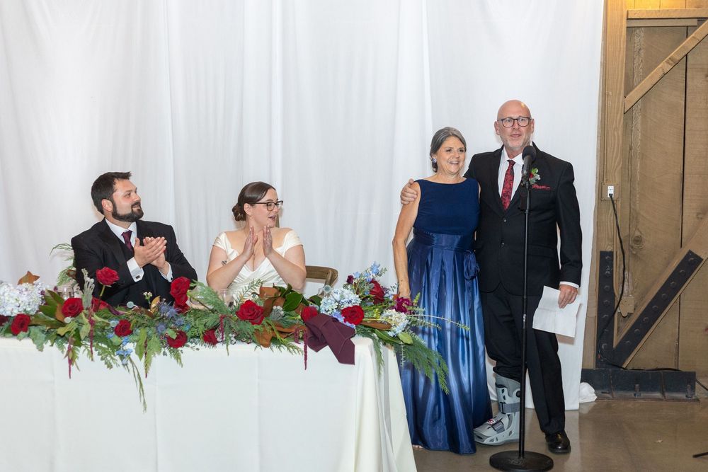 Wedding party members seated at a decorated head table with white draping and floral arrangements.