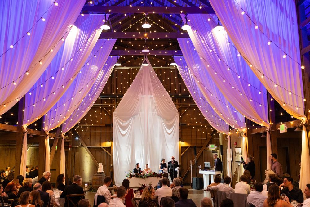 Wedding reception hall decorated with purple uplighting, white drapes, and string lights creating a magical atmosphere.