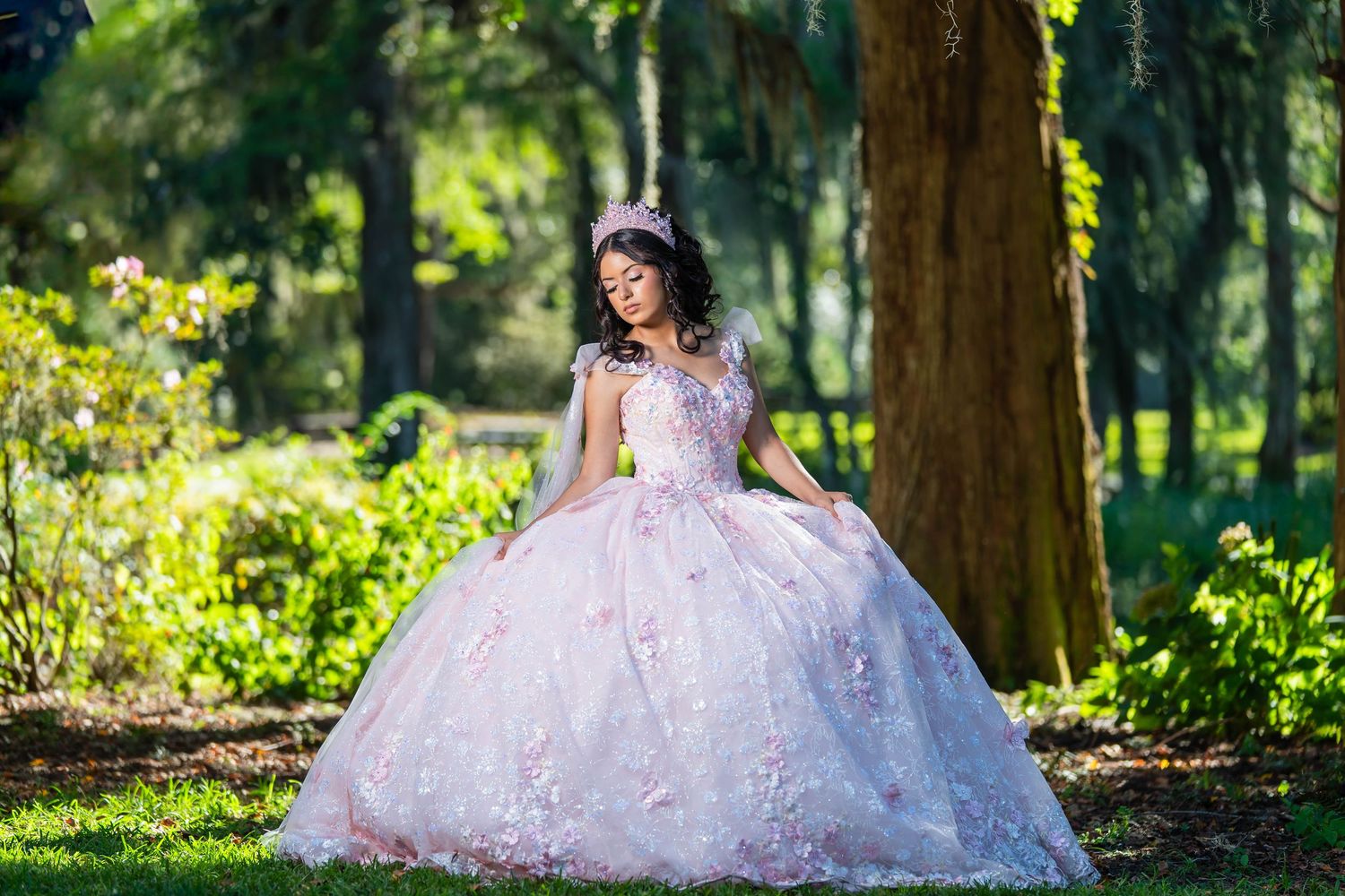 A person in a flowing white ballgown poses gracefully in a sunlit garden setting surrounded by lush greenery.