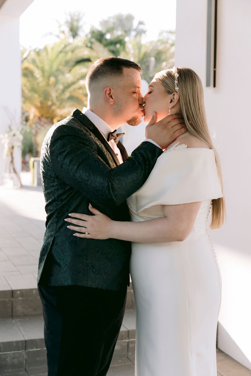 Romantic wedding couple embracing in white colonnade with palm trees.