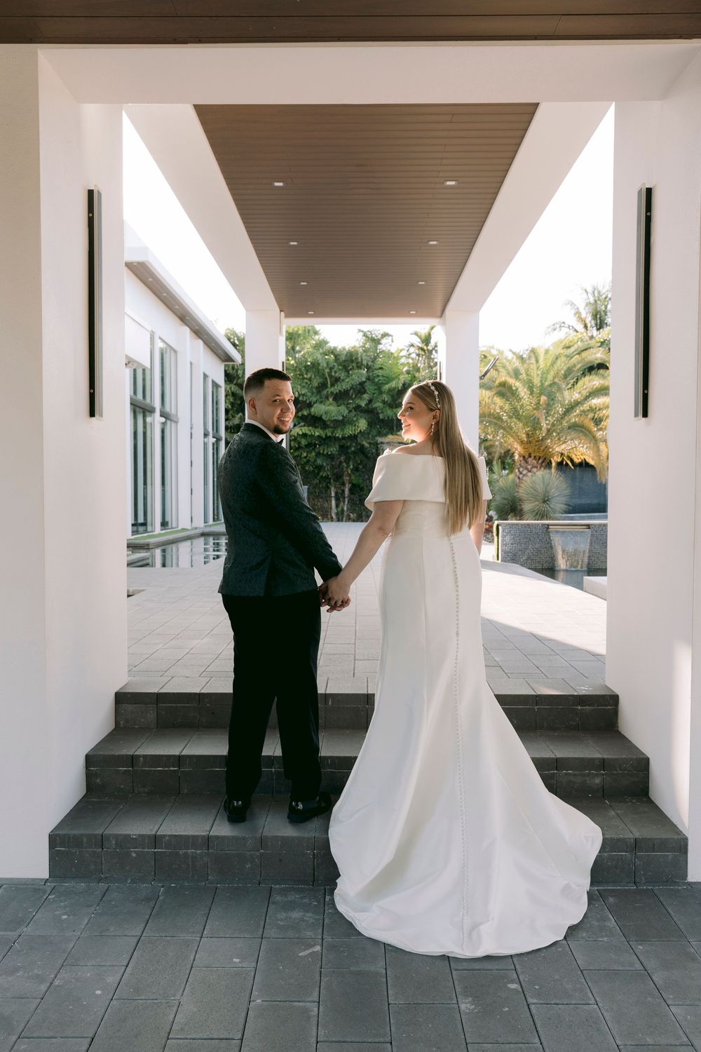 A wedding couple shares a tender moment in an elegant covered walkway with white columns and palm trees.