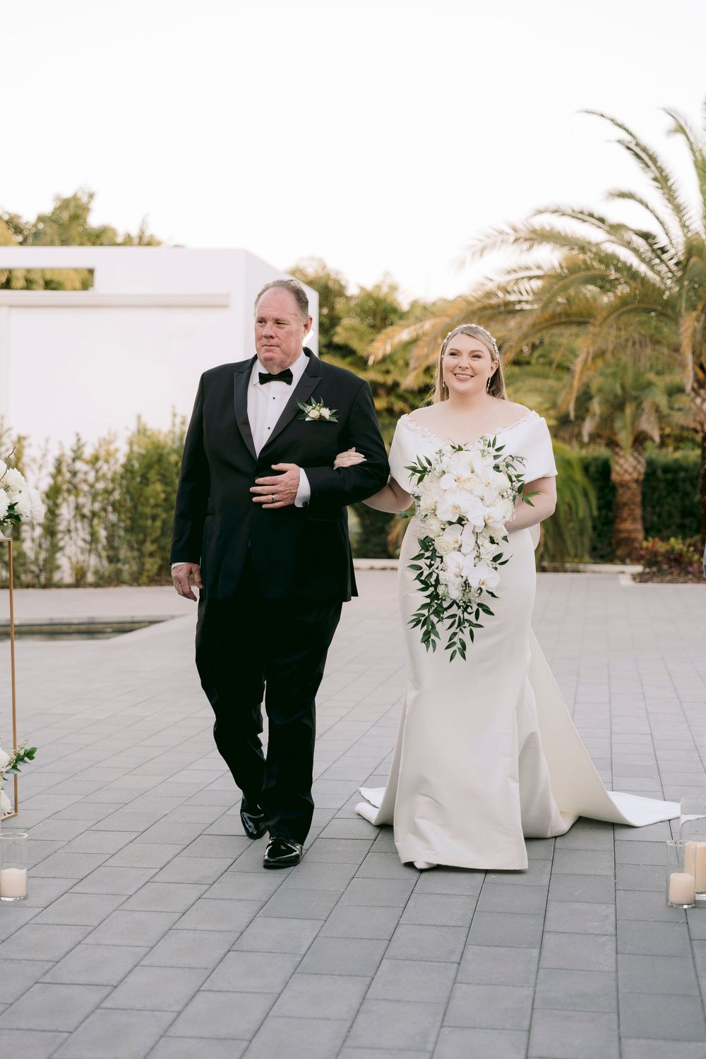 A wedding couple walks together on a modern stone terrace with palm trees and candles.