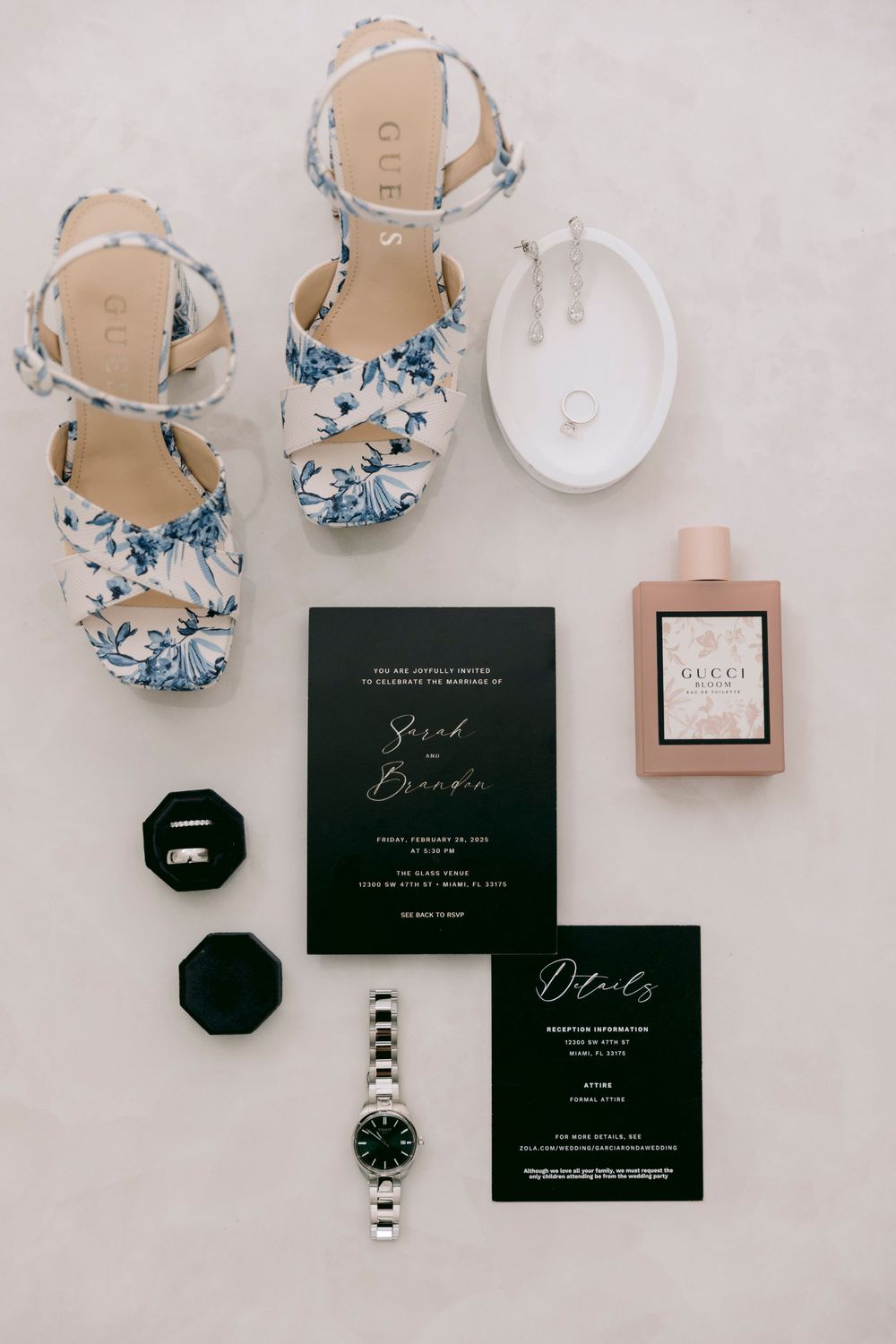 Wedding details flatlay with blue floral shoes, black invitations, perfume bottle, and watch on white surface.