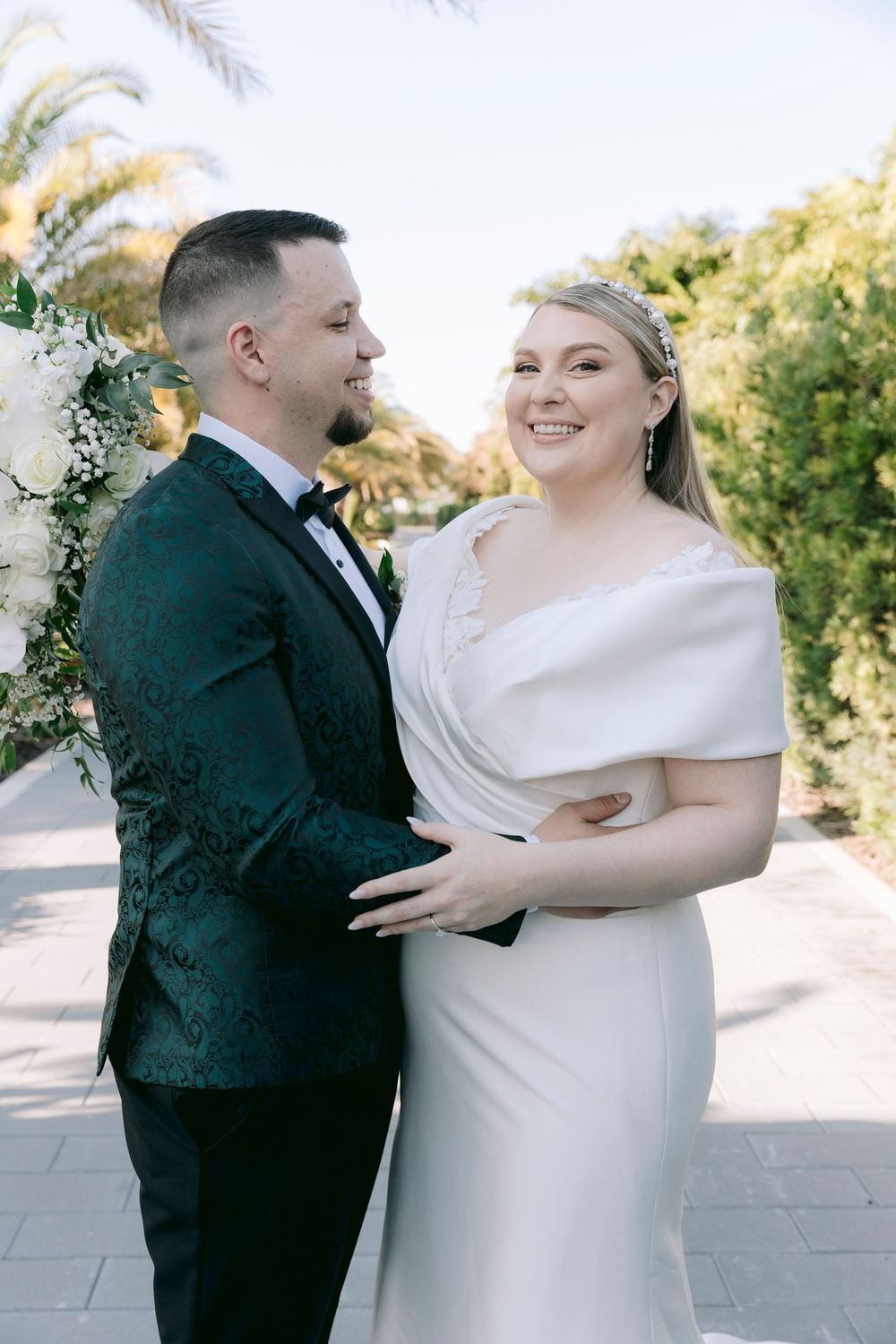 A joyful wedding couple shares laughter on an outdoor terrace with palm trees in the background.