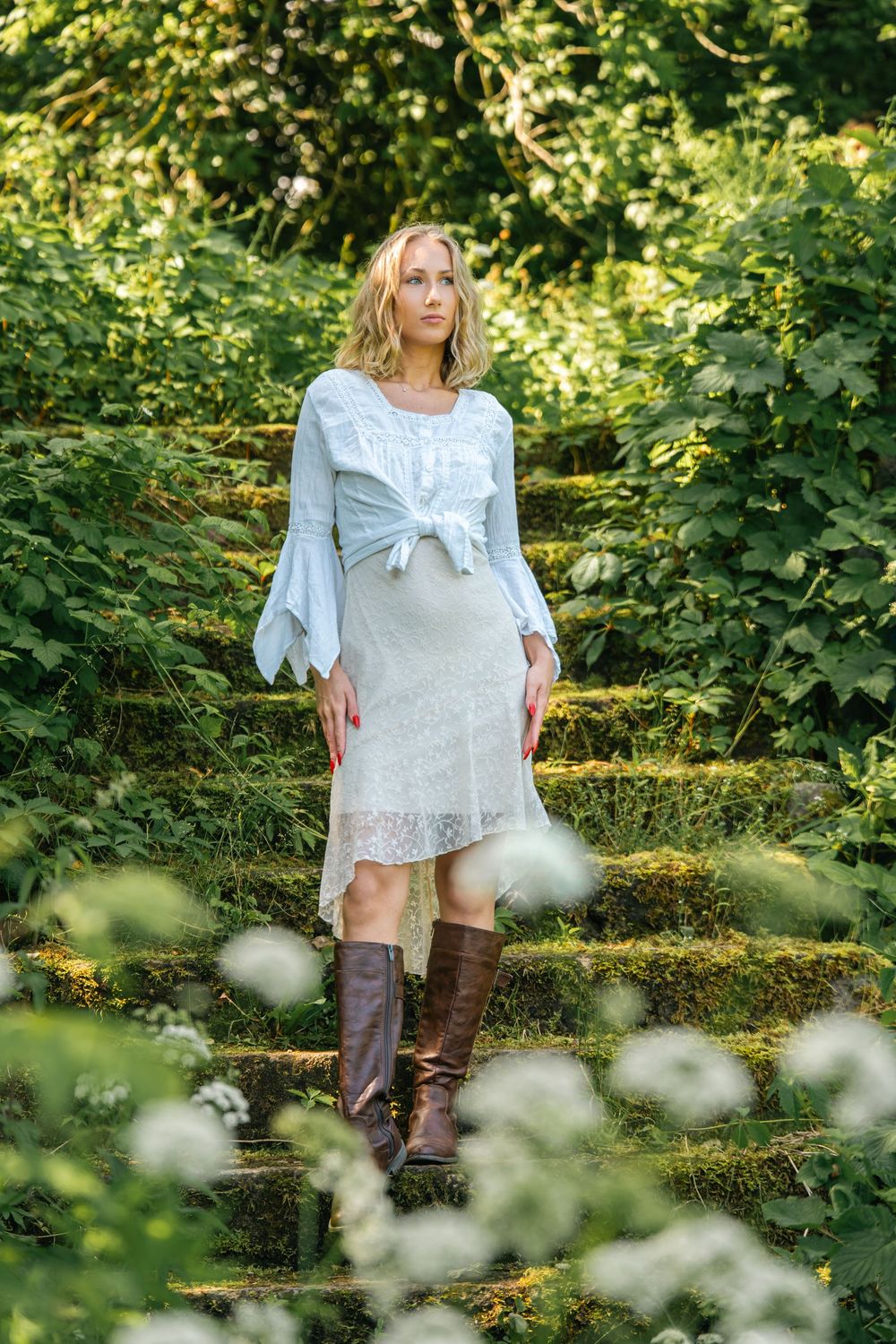 A white lace dress and brown boots create a romantic look on mossy garden steps surrounded by greenery.