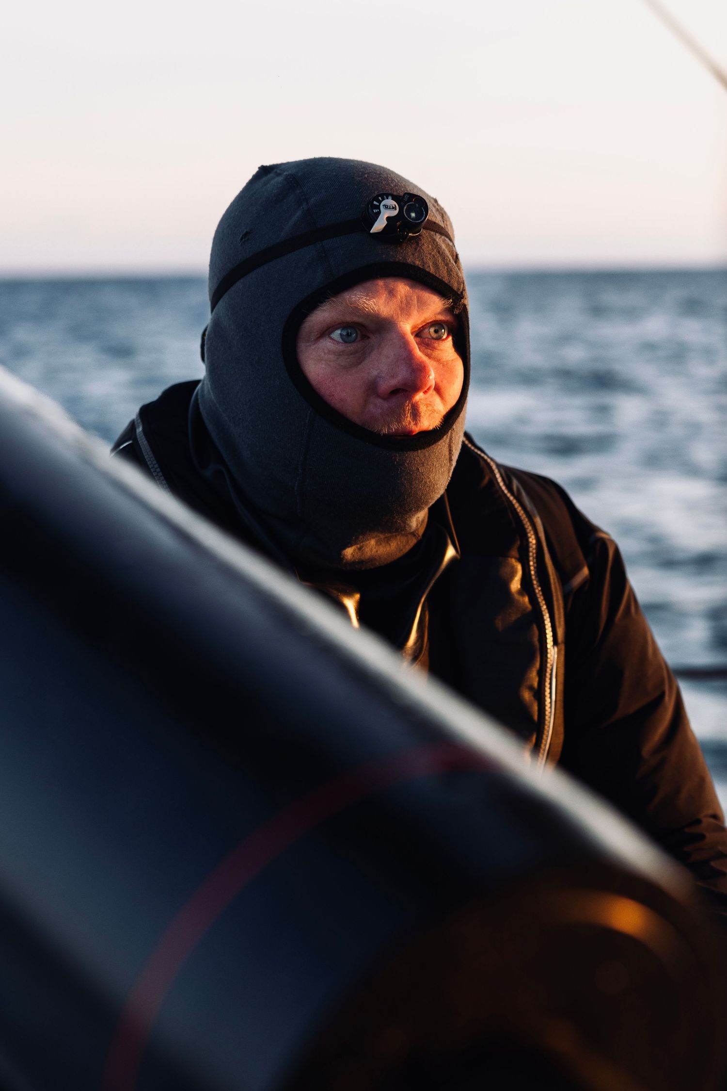 Sailor wearing dark winter gear steering a boat on choppy waters at dusk.
