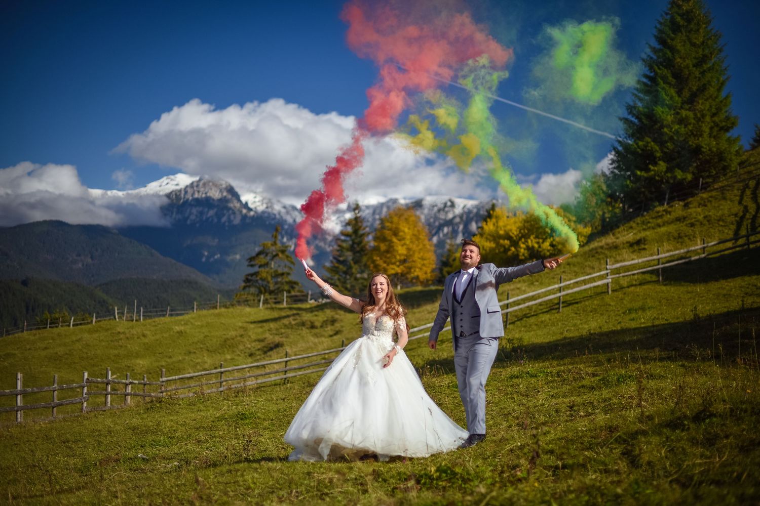 A couple holds colorful smoke flares on a scenic mountain meadow during their wedding photoshoot.