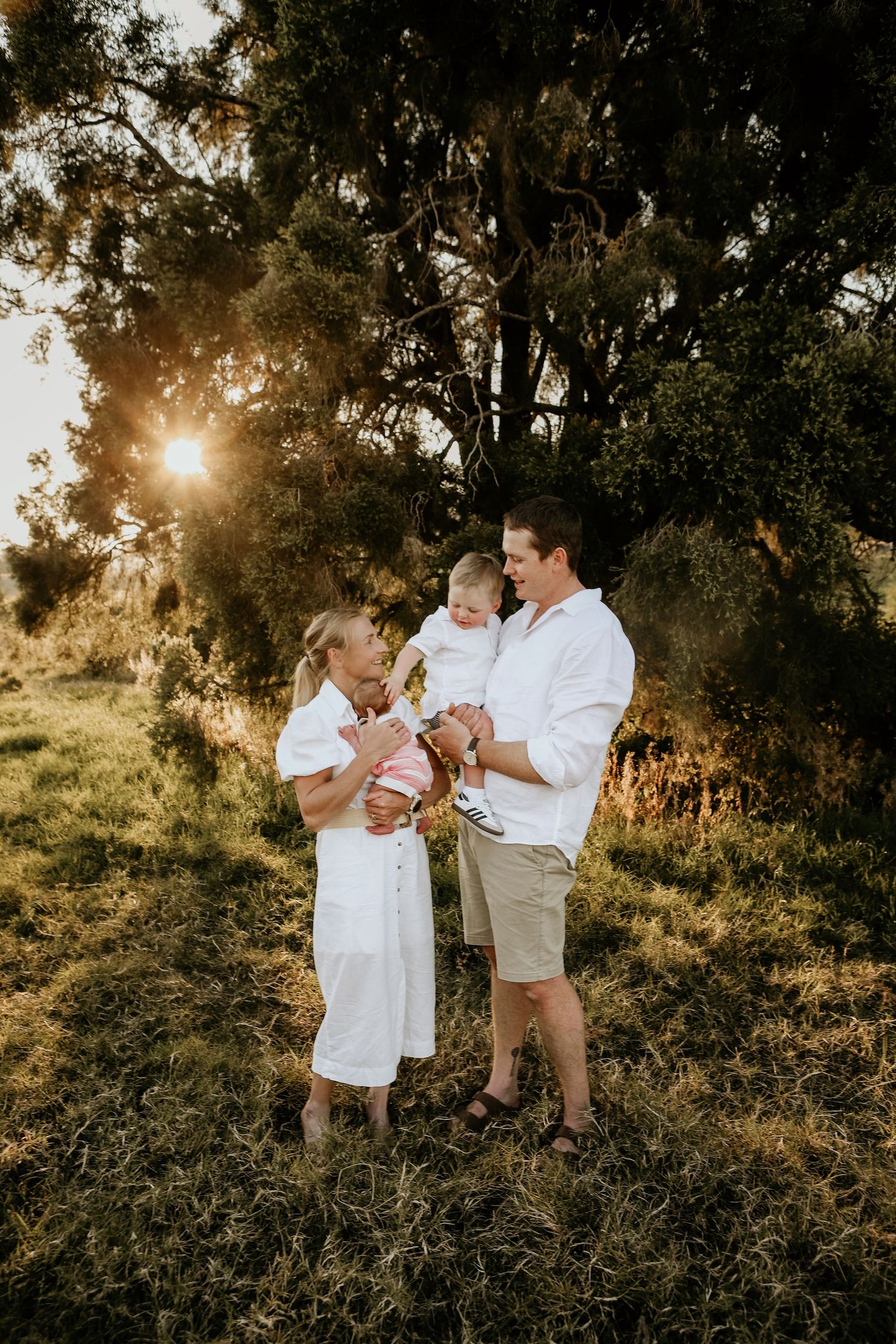 A family of three in white outfits poses together in a sunlit outdoor setting with trees at golden hour.