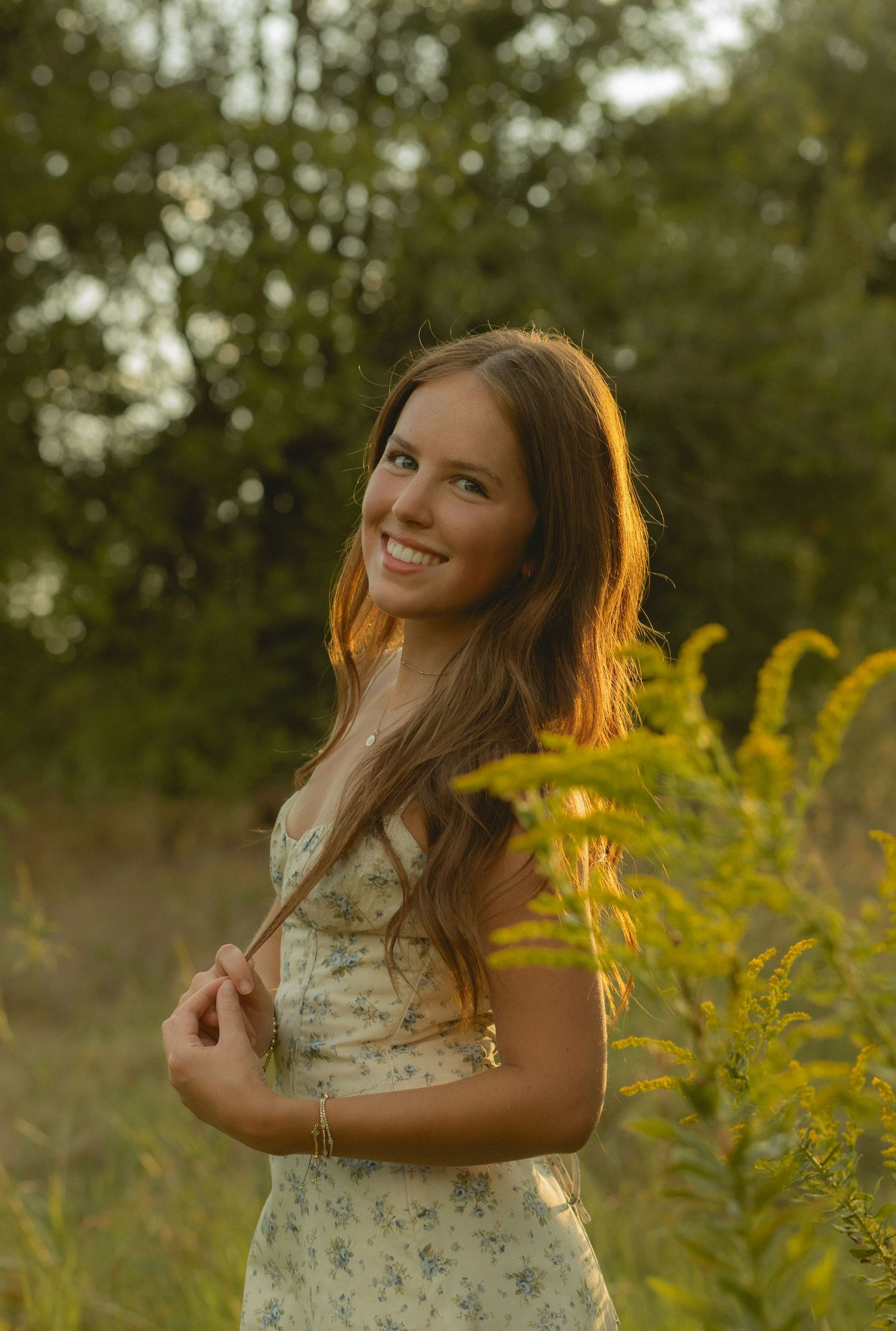 Smiling young woman in floral dress standing in golden hour sunlight among wildflowers and green trees.