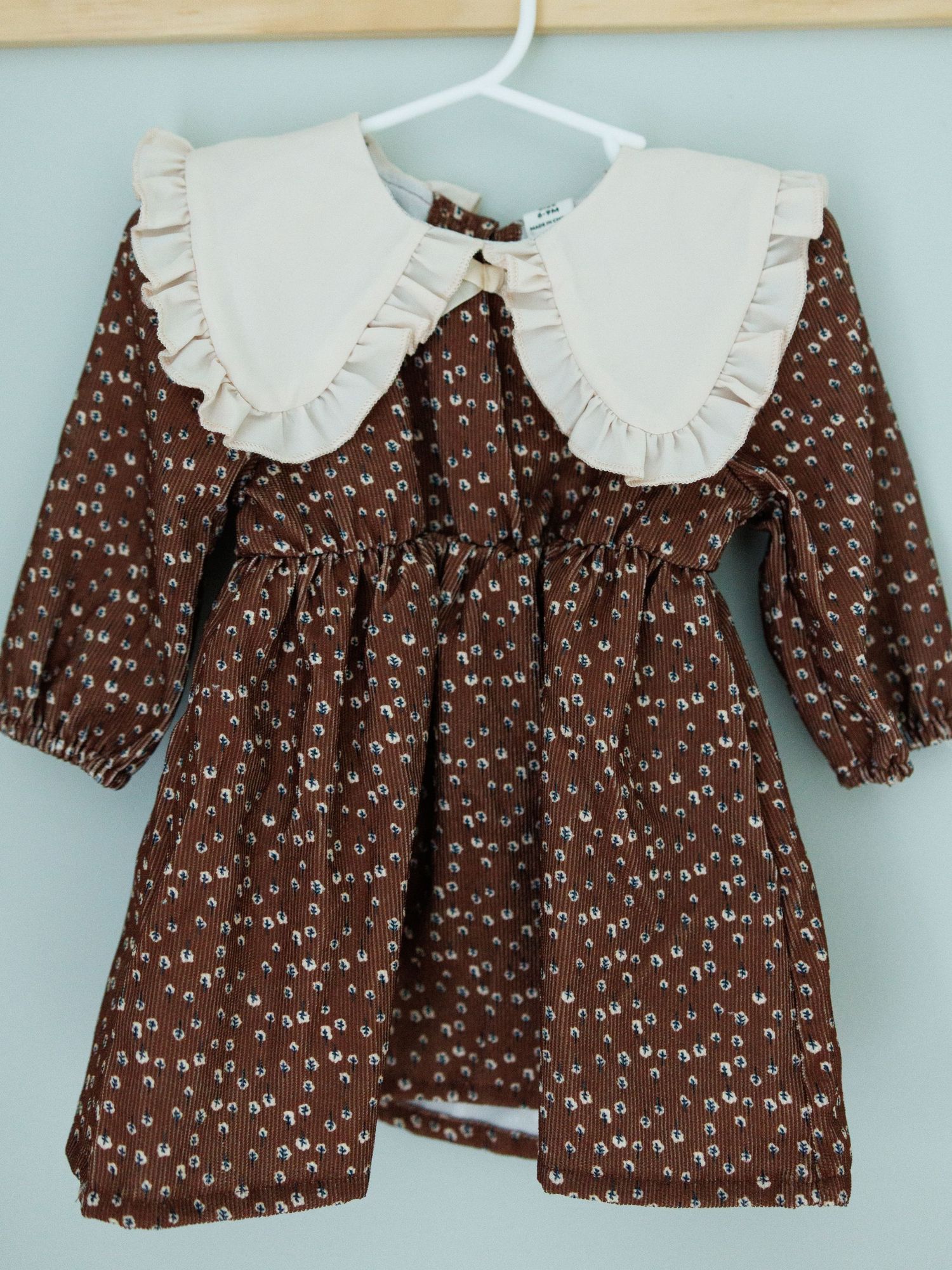 Formal infant outfit with beige vest, white shirt, light blue bow tie, and matching shorts hanging on wooden ladder shelf.