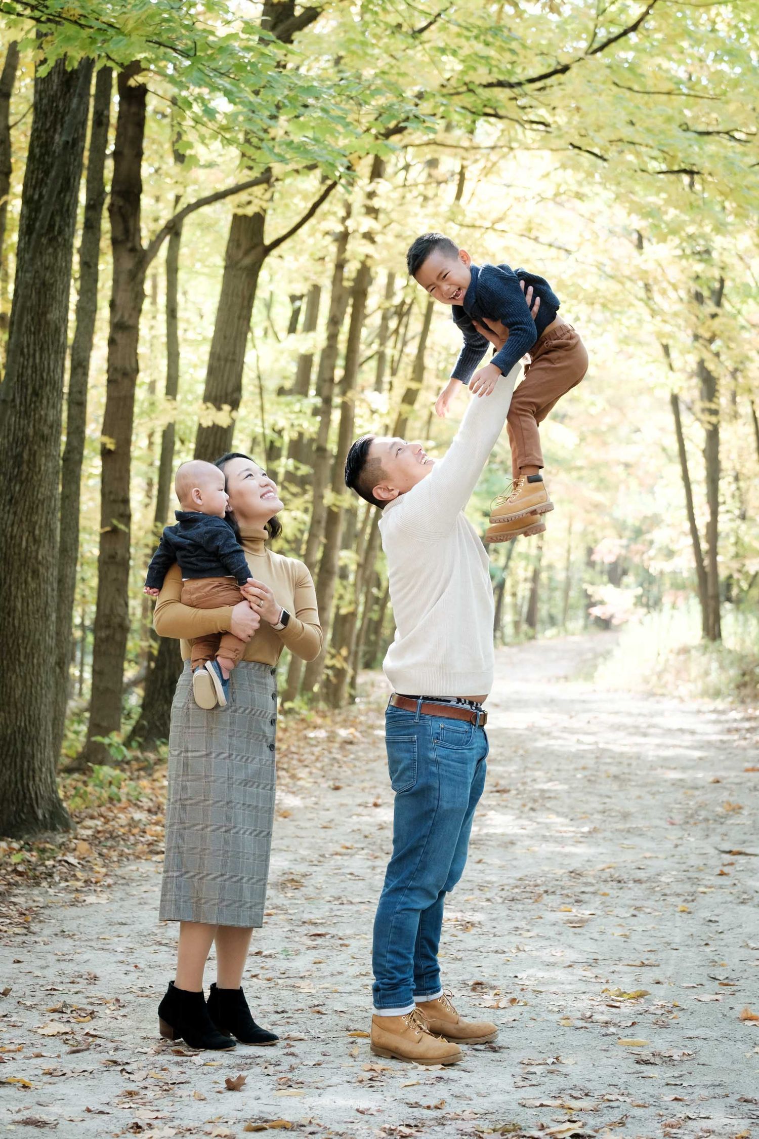 A family plays together on a tree-lined path covered in fallen autumn leaves.