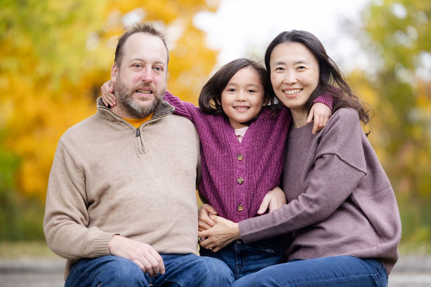 A family sits together outdoors during autumn with golden leaves in the background.