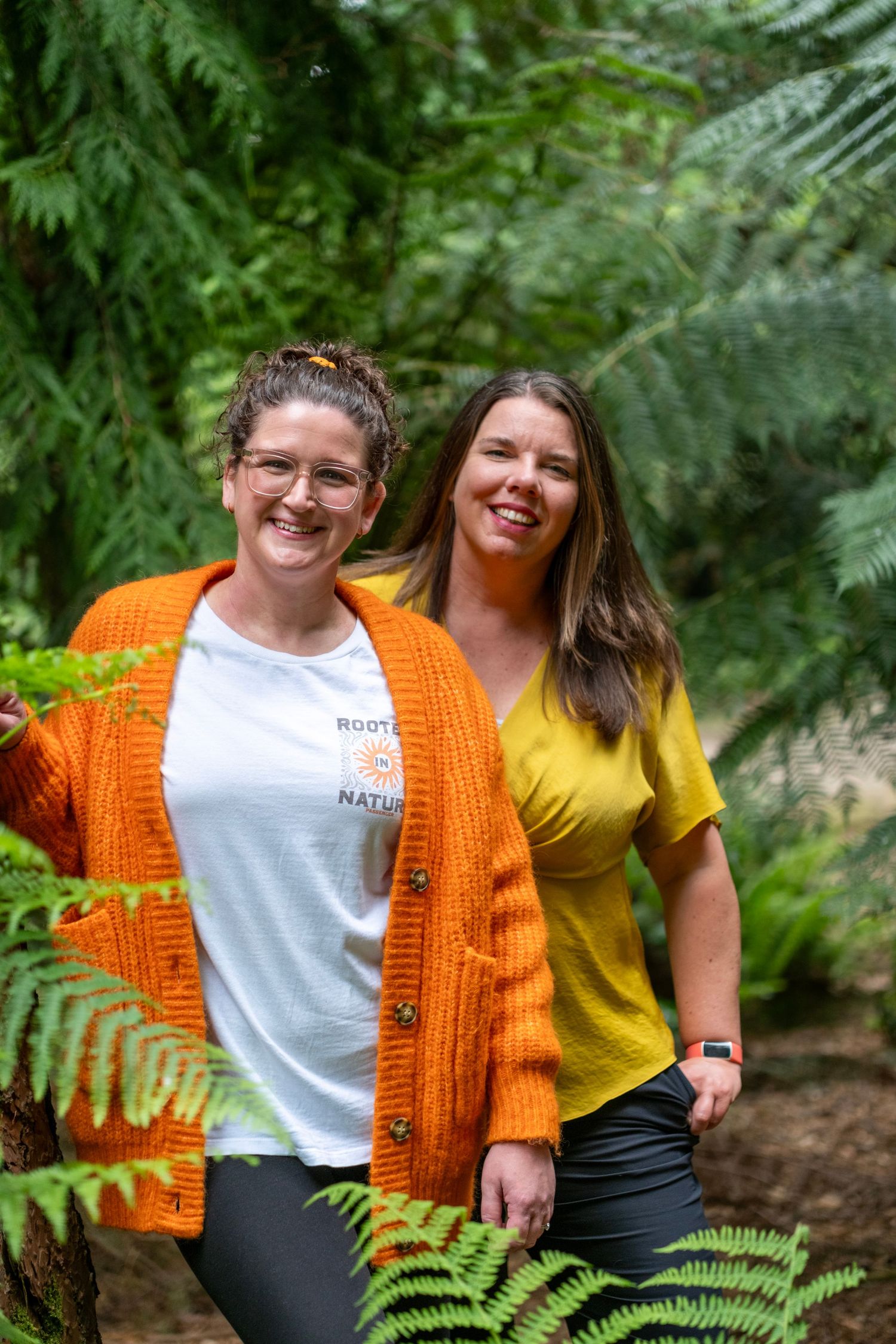 Two people wearing orange and yellow tops smile while standing among lush green ferns in an outdoor setting.