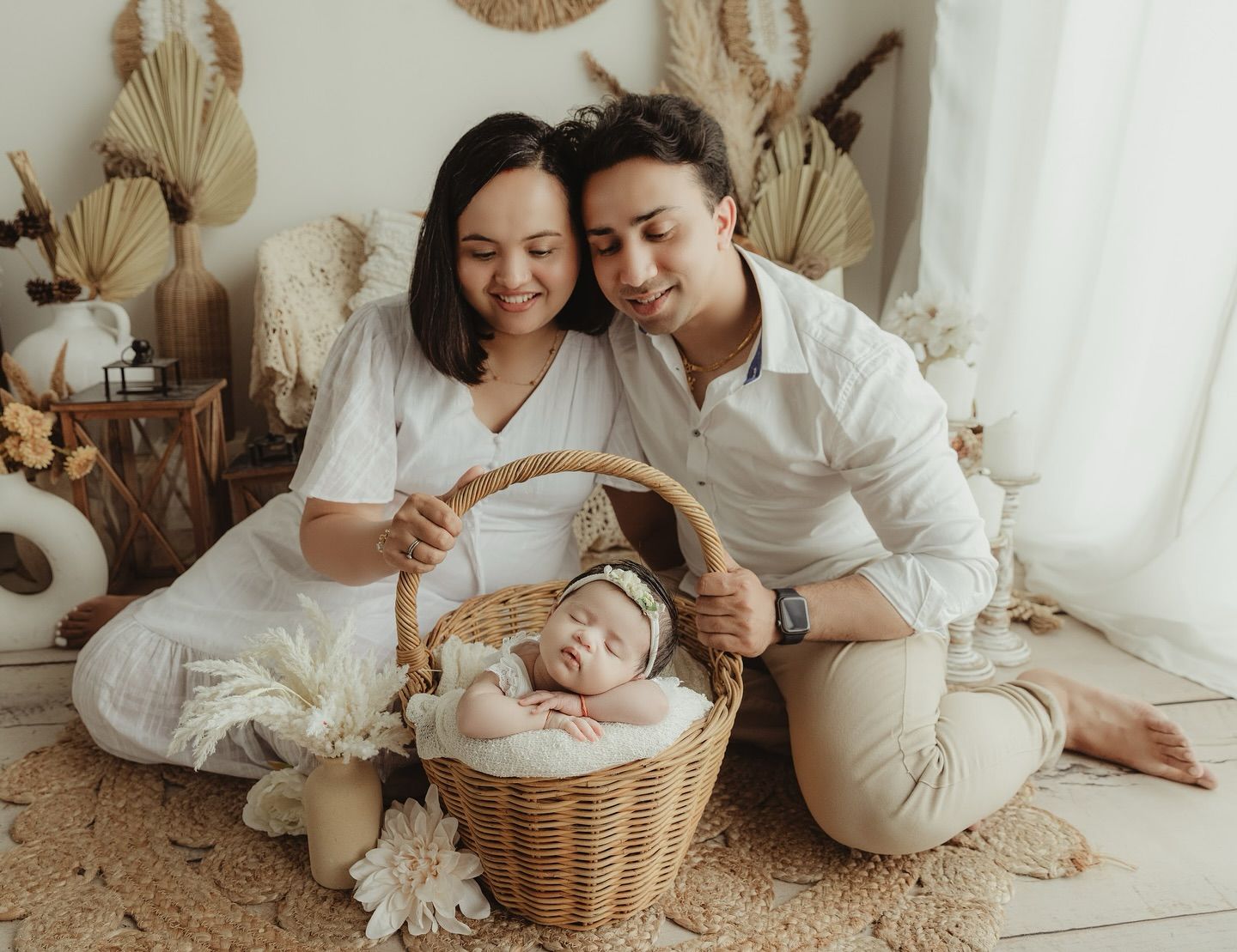 Family in white outfits relaxes together on floor with woven basket and neutral decor in soft studio setting.