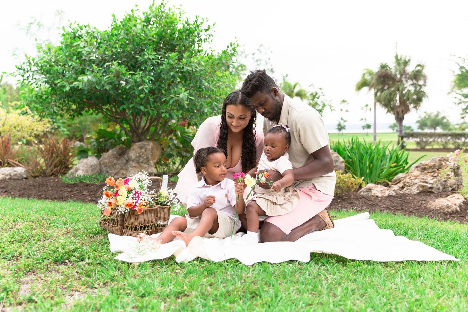 A joyful Black family of four enjoys a picnic on a white blanket in a lush green park setting.