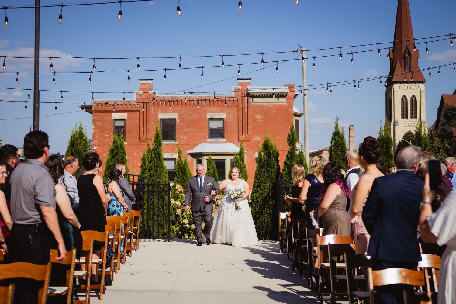 Outdoor wedding ceremony setup with string lights, brick building backdrop and rows of wooden chairs under blue sky.