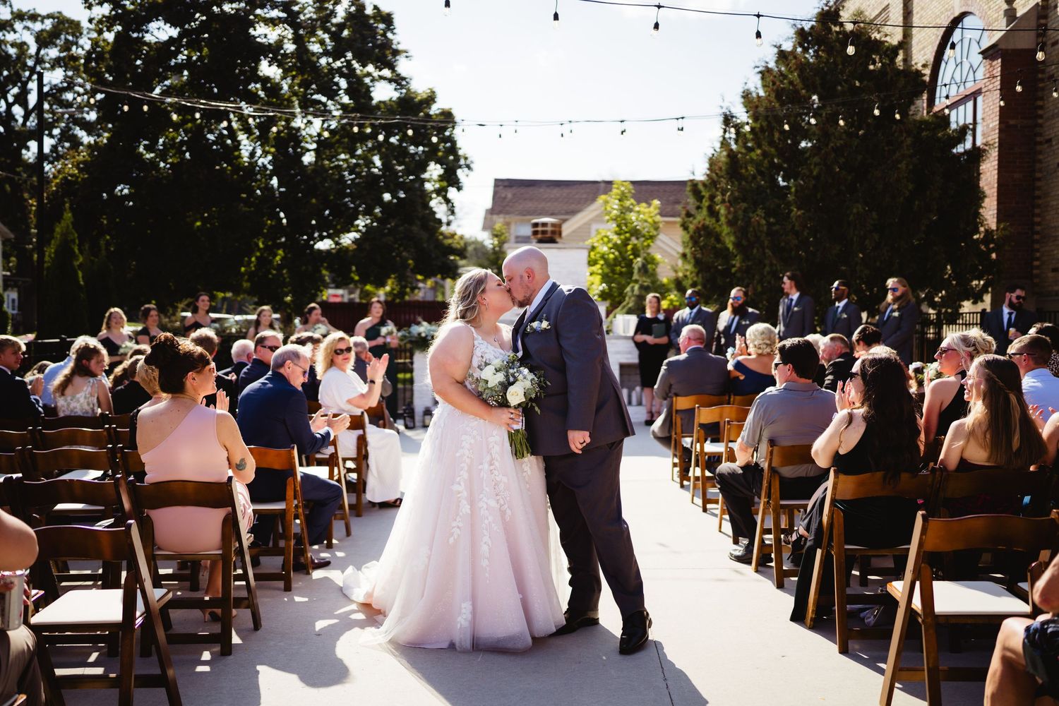 A wedding ceremony takes place outdoors with guests seated in wooden pews and string lights hanging overhead.