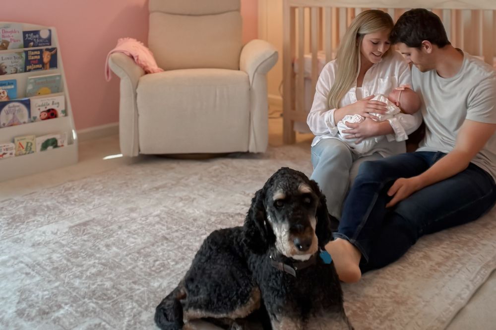 Young couple with newborn baby and dog sitting in a pink nursery room.