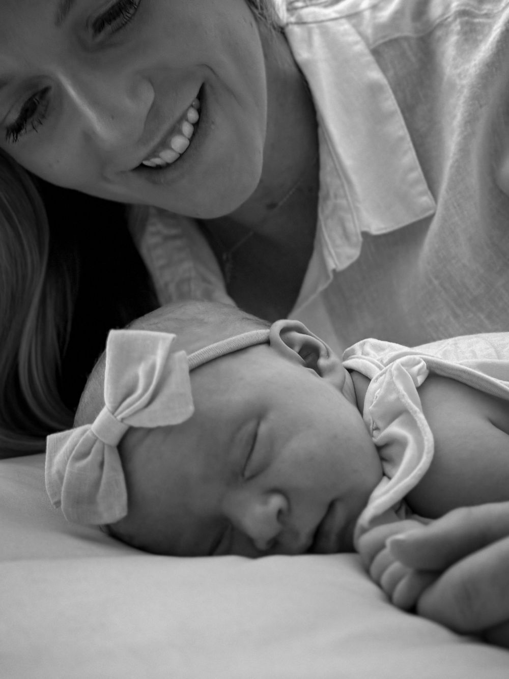 Black and white photo of a smiling mother gazing at her sleeping newborn with a bow headband.