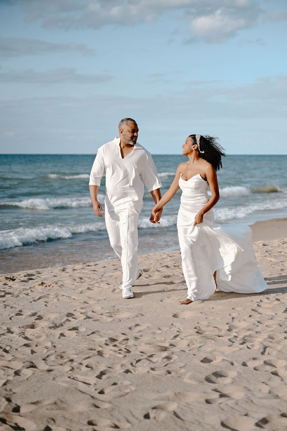 A couple in white outfits walks hand in hand along a sunny beach with ocean waves in the background.